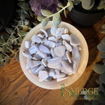 A bowl filled with polished blue lace agate tumbled stones, photographed from above on a wooden surface with greenery and a purple crystal in the background. The brand name 'SMUDGE' is visible as a watermark at the bottom right.