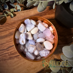A bowl filled with polished tumbled crystals blue lace agate small, set on a wooden surface with decorative leaves around it. A 'SMUDGE' brand logo is visible in the lower right corner.