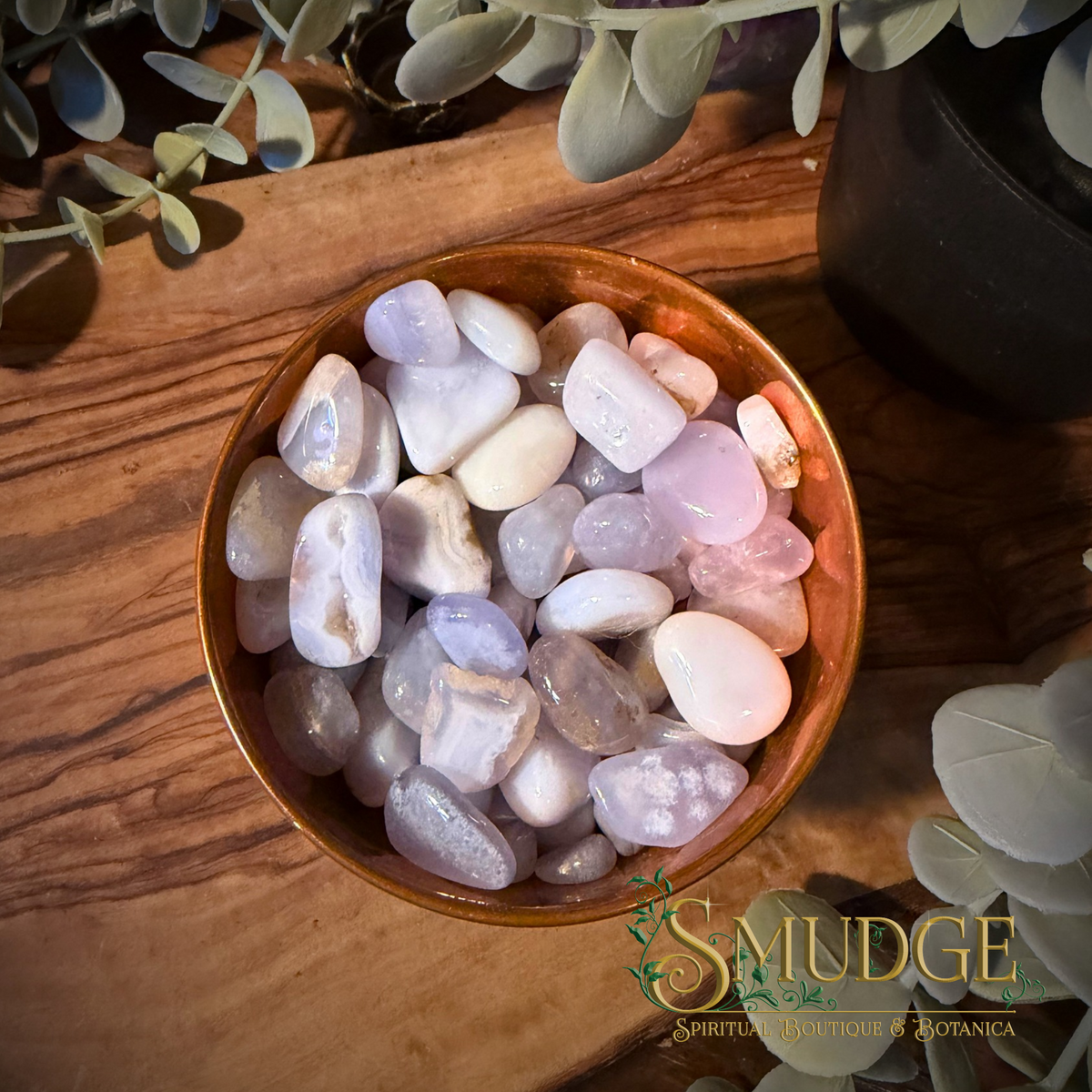 A bowl filled with polished tumbled crystals blue lace agate small, set on a wooden surface with decorative leaves around it. A 'SMUDGE' brand logo is visible in the lower right corner.