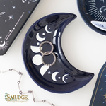 A glossy crescent moon-shaped jewelry tray in deep navy blue with white moon and star illustrations, holding two silver rings. The tray is styled on a white surface with other celestial-themed jewelry items and packaging boxes.