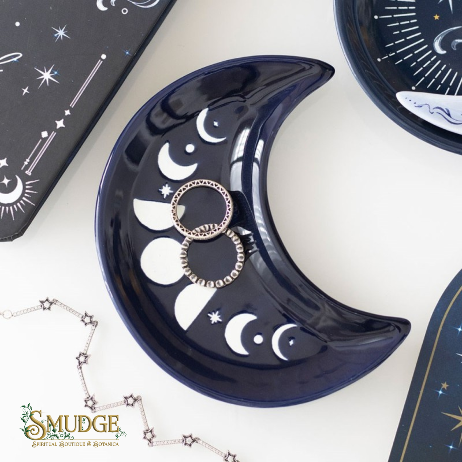 A glossy crescent moon-shaped jewelry tray in deep navy blue with white moon and star illustrations, holding two silver rings. The tray is styled on a white surface with other celestial-themed jewelry items and packaging boxes.