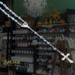 A silver rosary necklace with light blue faceted beads and a detailed crucifix pendant, displayed diagonally across a dark, atmospheric apothecary shop background with shelves of bottles and a chandelier.
