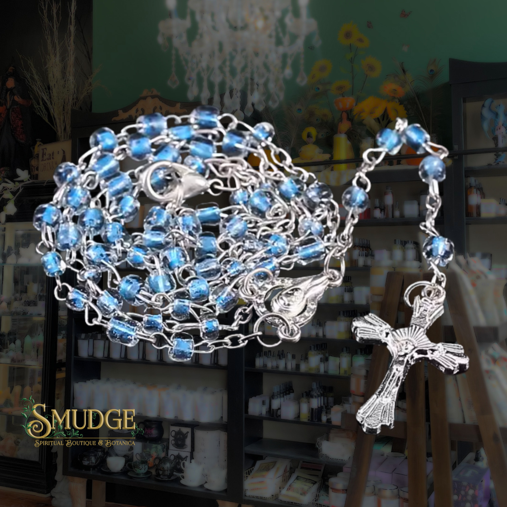 A silver-tone rosary necklace with light blue faceted beads and a detailed crucifix pendant, displayed in front of a blurred shop interior with shelves and decor. The word 'SMUDGE' appears as a watermark in the lower-left corner.