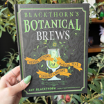 A hand holds a hardcover book titled "Blackthorn's Botanical Brews" against a background of green foliage. The book cover is dark gray with green and white lettering and an illustration of a potion glass.