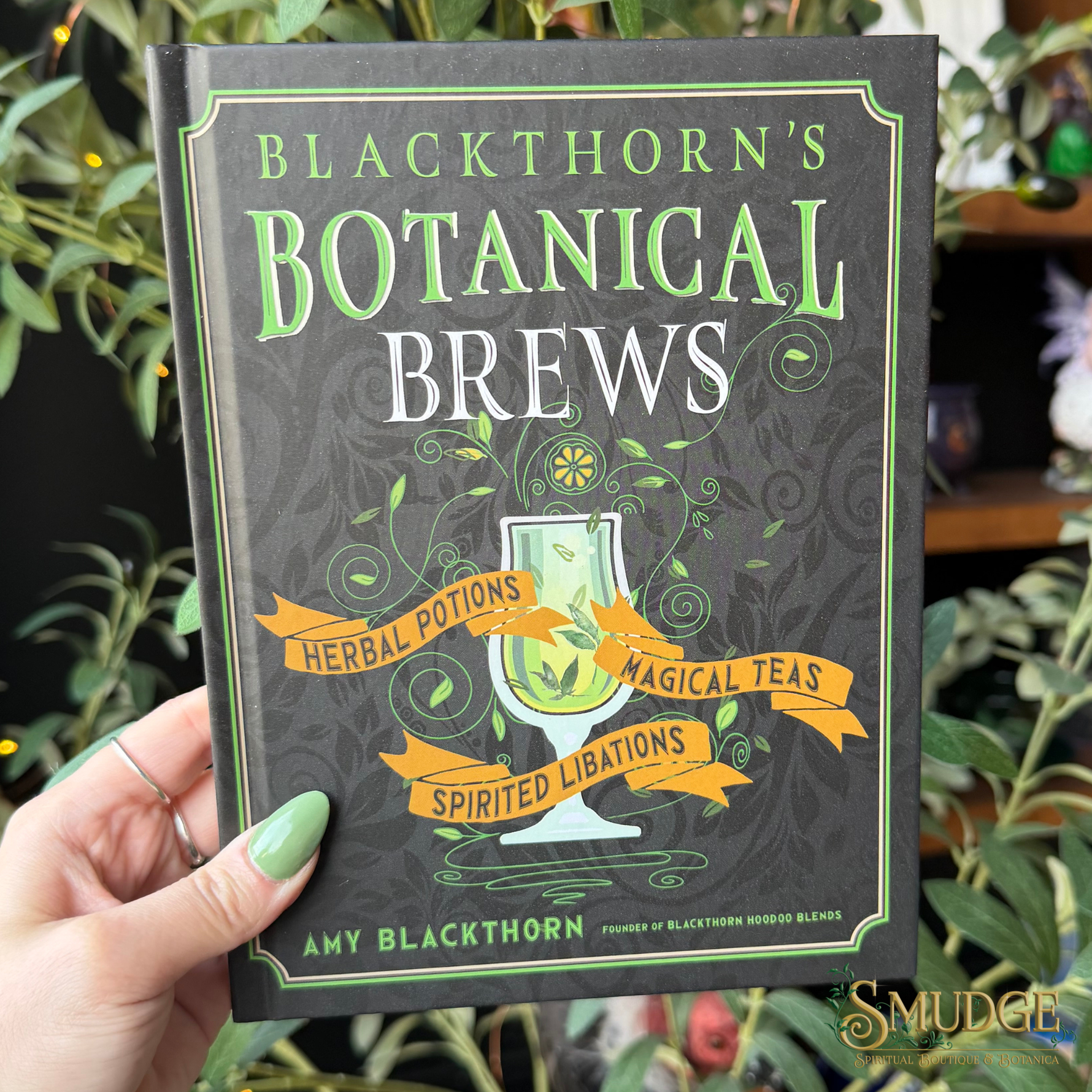 A hand holds a hardcover book titled "Blackthorn's Botanical Brews" against a background of green foliage. The book cover is dark gray with green and white lettering and an illustration of a potion glass.