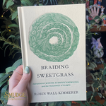 Book titled 'Braiding Sweetgrass' held by a hand with green nail polish, surrounded by plants.