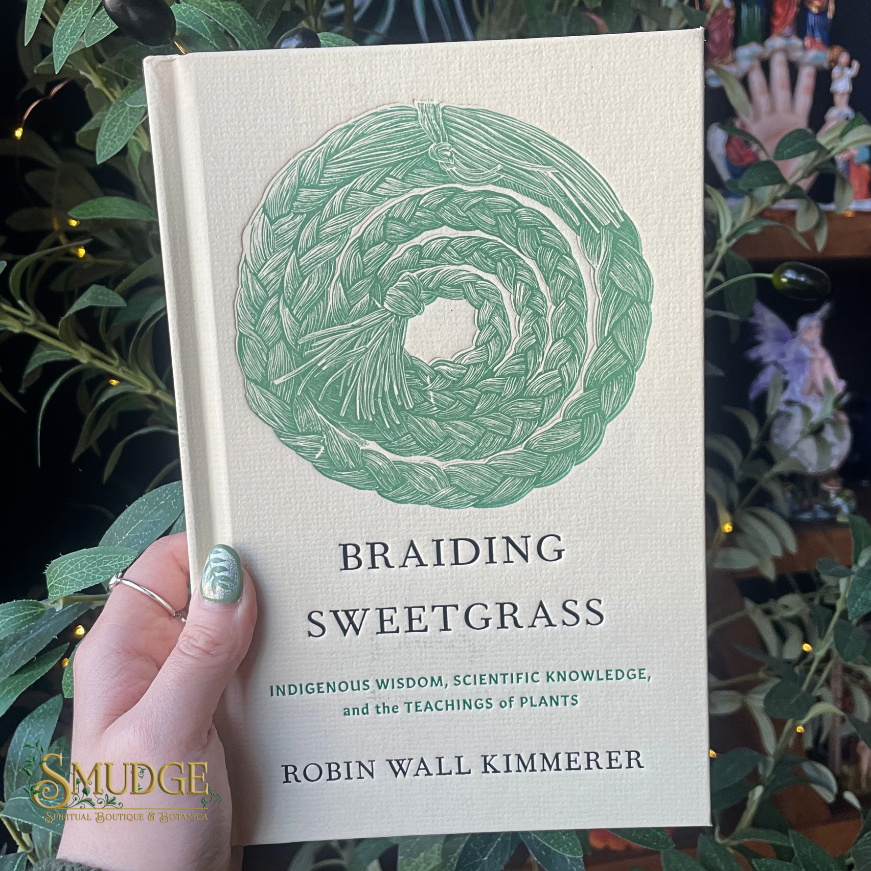 Book titled 'Braiding Sweetgrass' held by a hand with green nail polish, surrounded by plants.