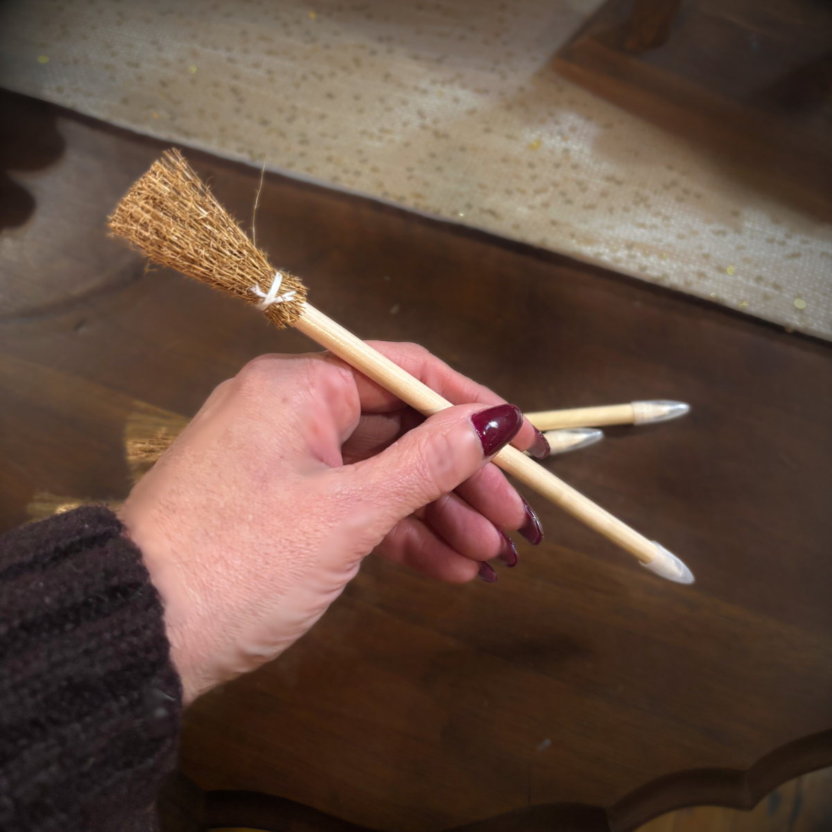 Hand holding a small broom with a wooden handle and bristle head on a reflective surface.