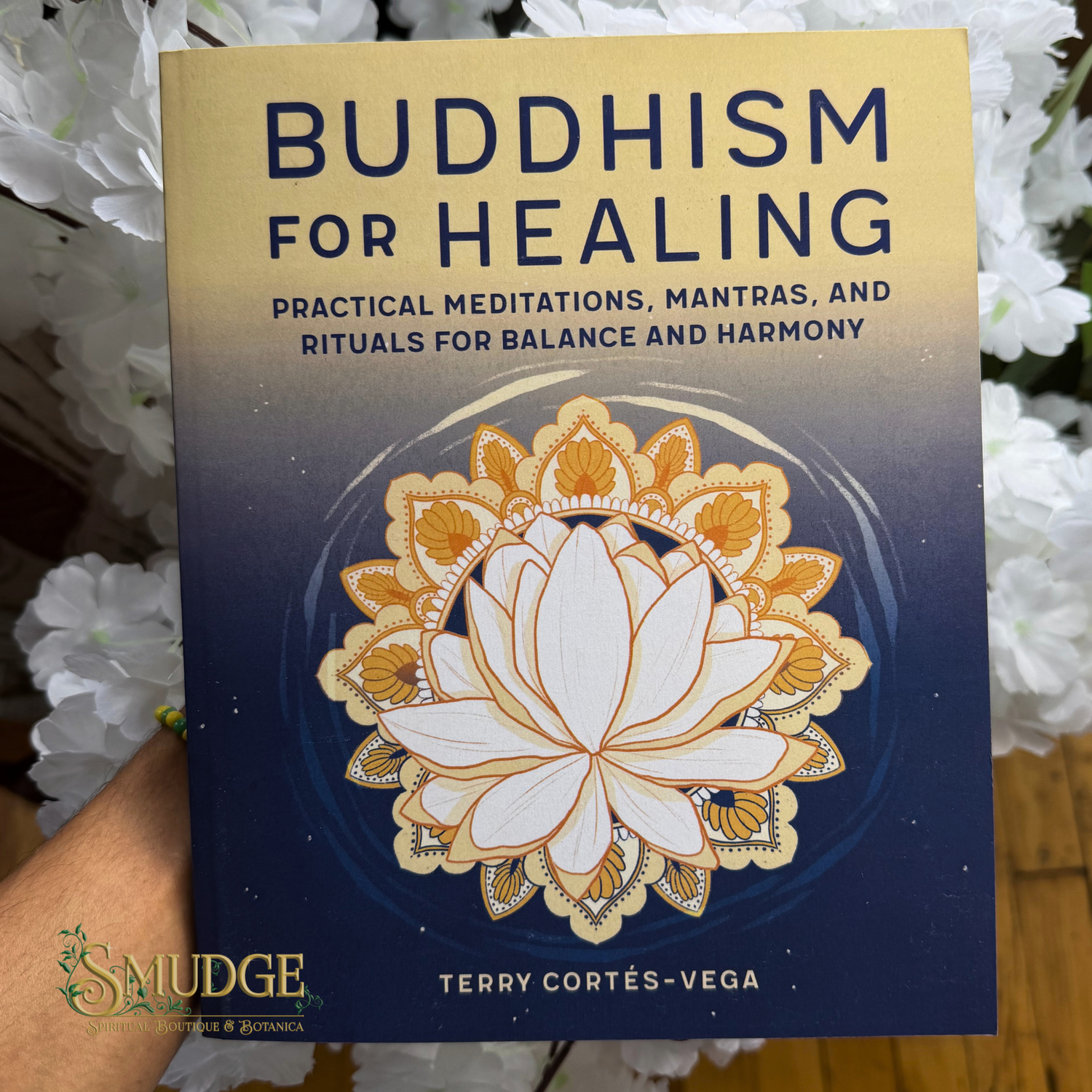 The front cover of the book 'Buddhism for Healing' by Terry Cortés-Vega is shown upright, held by a hand with white flowers in the background. The cover features a gold-to-dark-blue gradient with a large white and gold lotus mandala in the center.