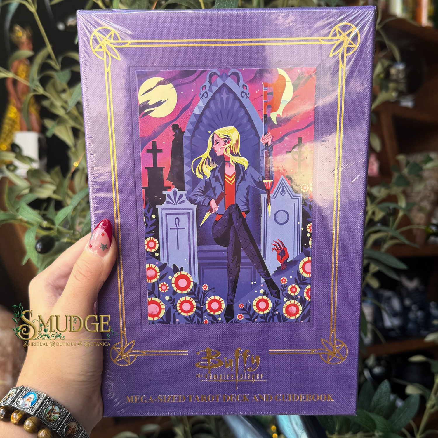 A hand holds a shrink-wrapped purple tarot deck box with an illustrated cover featuring a blonde woman in a graveyard scene. The box has gold decorative border lines and text reading 'Buffy the Vampire Slayer' and 'Mega Sized Tarot Deck and Guidebook.