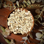 A bowl filled with dried, chopped herbal roots or bark pieces, styled on a dark wooden surface with decorative leaves and flowers. The brand logo 'The Mudge Shop' is visible in the upper right.
