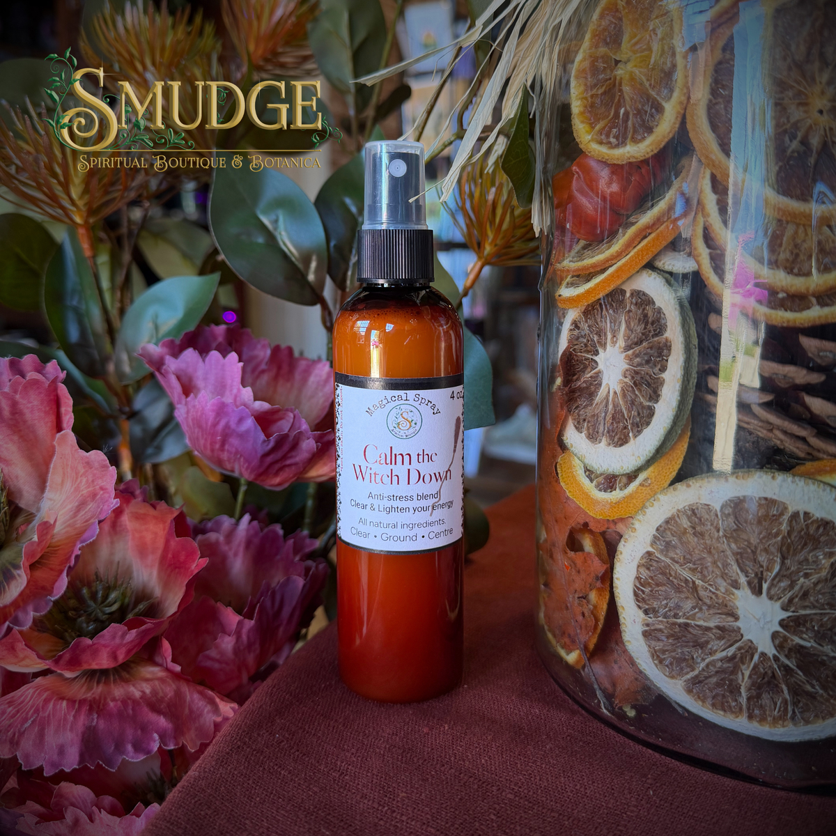 A bottle of Smudge Spirituale Boutiques & Botanicals 'Calm the Witch Down' anti-itch body spray, set on a dark red cloth next to a large glass jar filled with dried citrus slices and botanicals, with pink flowers in the background.
