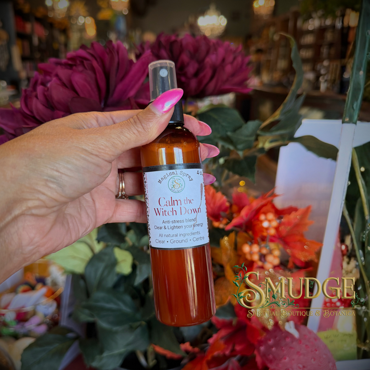 A hand with pink nail polish holds a bottle of 'Calm the Witch Down' anti-stress herbal spray in front of a blurred background of flowers and a shop interior. The brand 'Smudge Spiritual Goods' is visible as a watermark in the bottom right corner.
