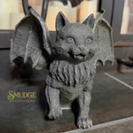 A gray cast-iron gargoyle figurine of a winged cat with a textured, weathered finish, standing on a dark surface. A 'SMUDGE' brand logo is visible in the lower-left corner.