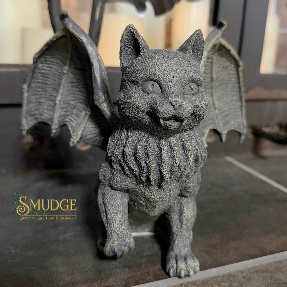 A gray cast-iron gargoyle figurine of a winged cat with a textured, weathered finish, standing on a dark surface. A 'SMUDGE' brand logo is visible in the lower-left corner.