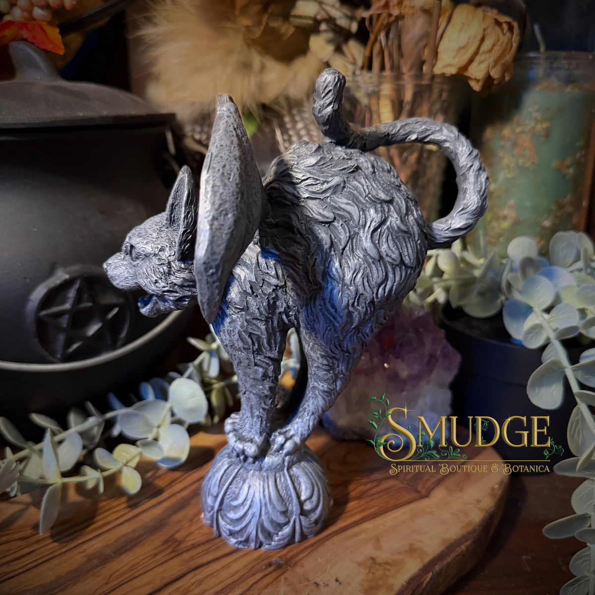 A gray cat-shaped spiritual statue with wings and a carved base, displayed on a wooden surface with dried flowers and decorative plants in the background. A 'SMUDGE Spiritual Botanicals' logo is visible on the right.