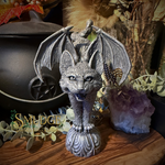 A silver bat-headed cat statue with spread wings, standing on a decorative base, displayed on a wooden surface with dried botanicals, a black cauldron, and an amethyst cluster in the background.