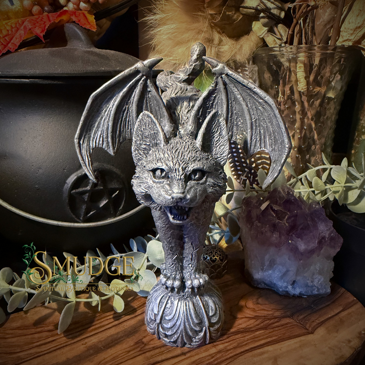 A silver bat-headed cat statue with spread wings, standing on a decorative base, displayed on a wooden surface with dried botanicals, a black cauldron, and an amethyst cluster in the background.