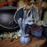 A gothic-style bat figurine with wings spread, standing on a carved pedestal. The bat is detailed with textured gray and silver tones.
