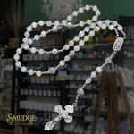 A rosary necklace with white pearl-like beads and silver-toned metal links, featuring a detailed crucifix pendant, displayed in front of a blurred shop interior. The SMUDGE logo is visible in the bottom-left corner.