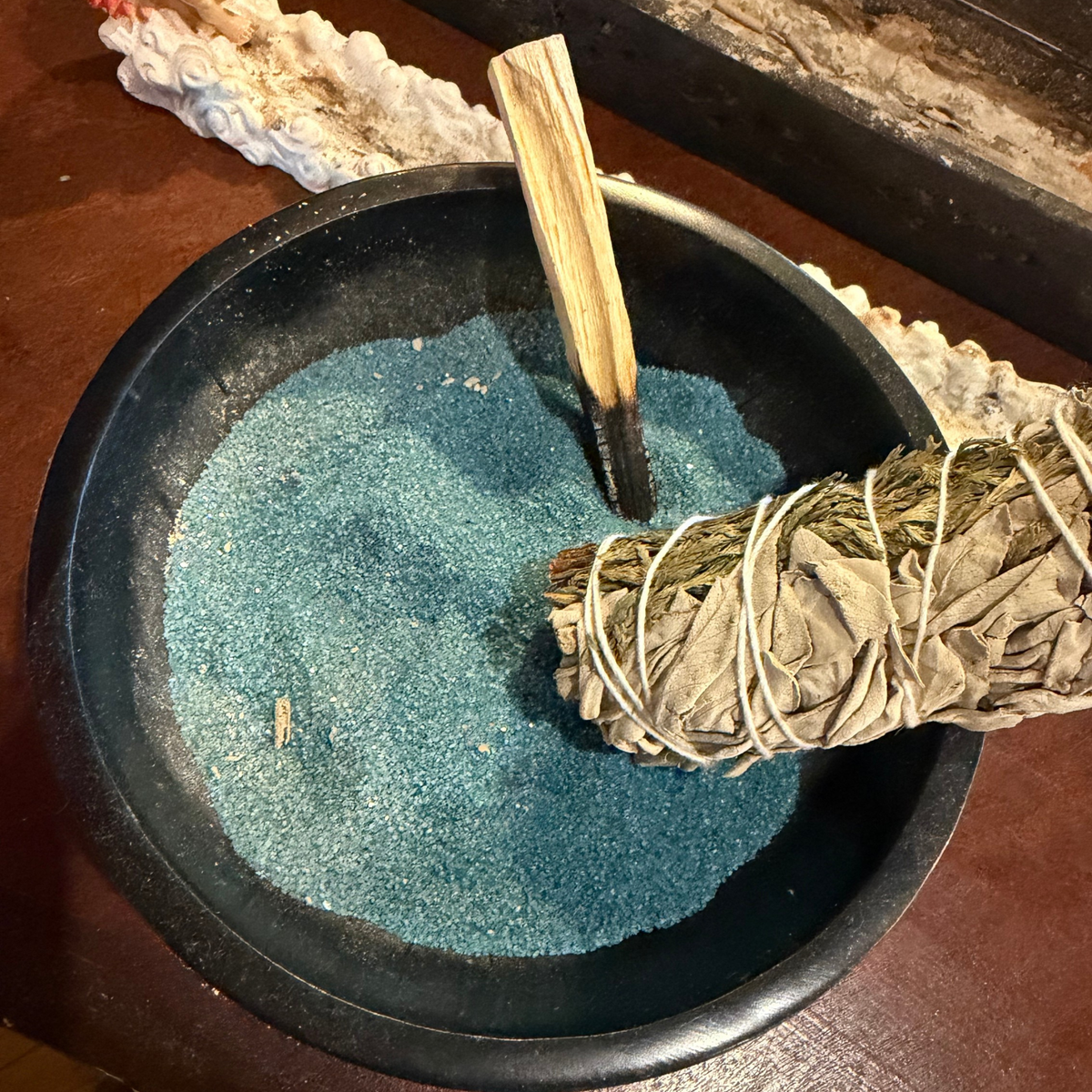 black bowl with blue coloured incense sand in it with a sage stick and palo santo. 