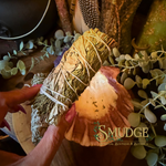 A hand holds a bundle of white sage and cedar tied with white string, alongside a sliced piece showing the inner spore-bearing structure, in a styled setting with eucalyptus leaves and a wooden surface. The brand name 'SMUDGE' is visible as an overlay.