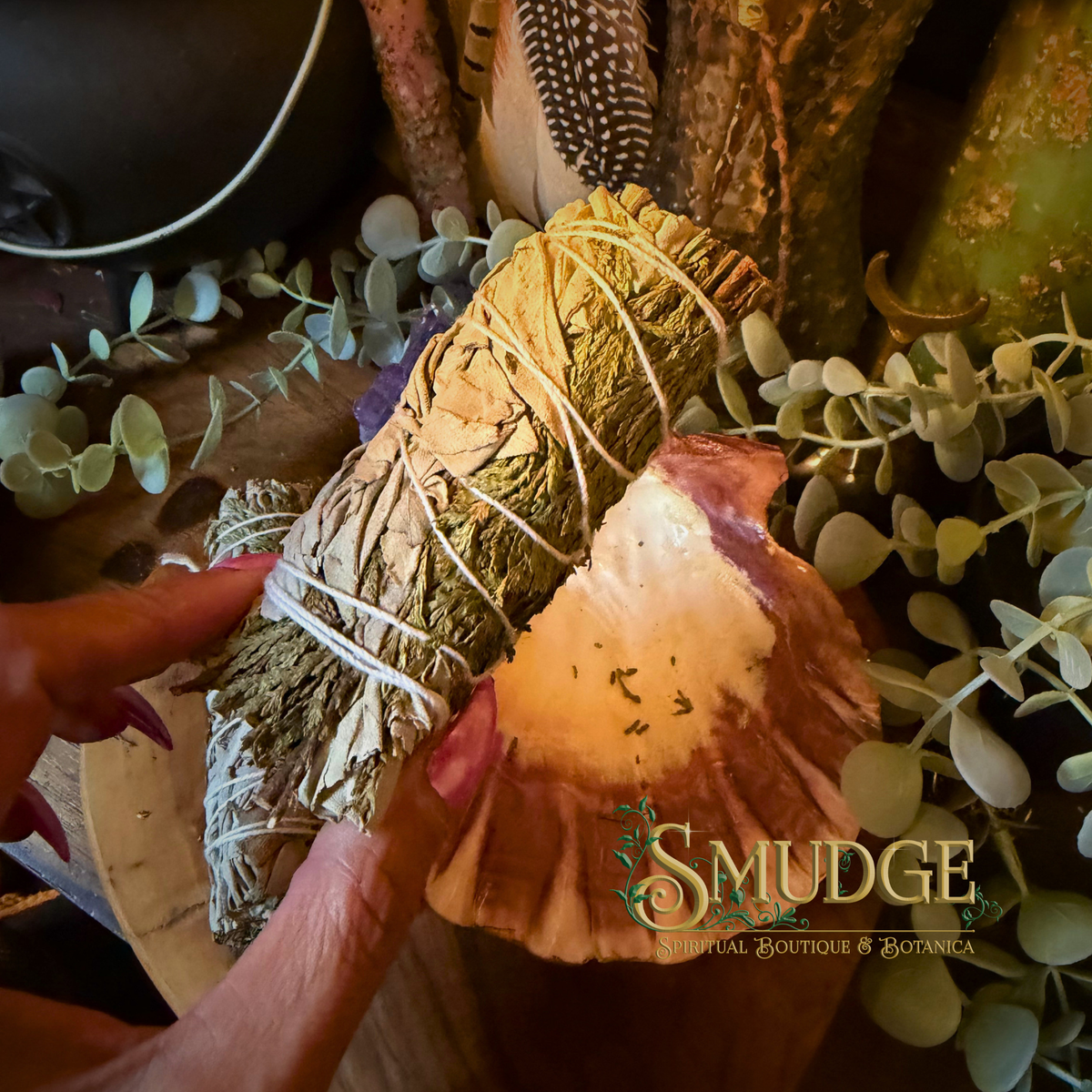 A hand holds a bundle of white sage and cedar tied with white string, alongside a sliced piece showing the inner spore-bearing structure, in a styled setting with eucalyptus leaves and a wooden surface. The brand name 'SMUDGE' is visible as an overlay.