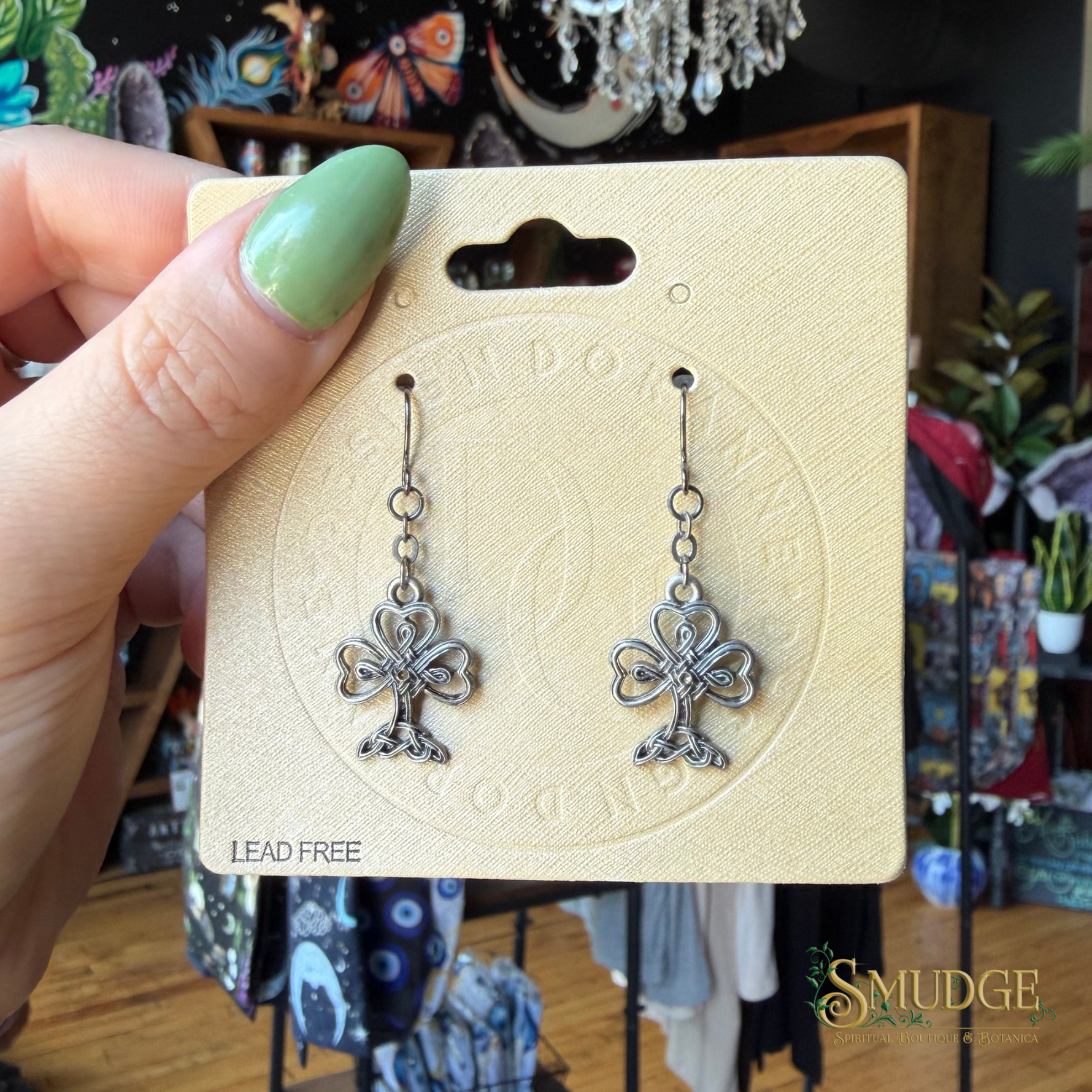 A hand with green nail polish holds a display card featuring a pair of dangling earrings. The earrings are silver-toned with a floral or clover-like design and are mounted on a textured beige card labeled 'LEAD FREE'.