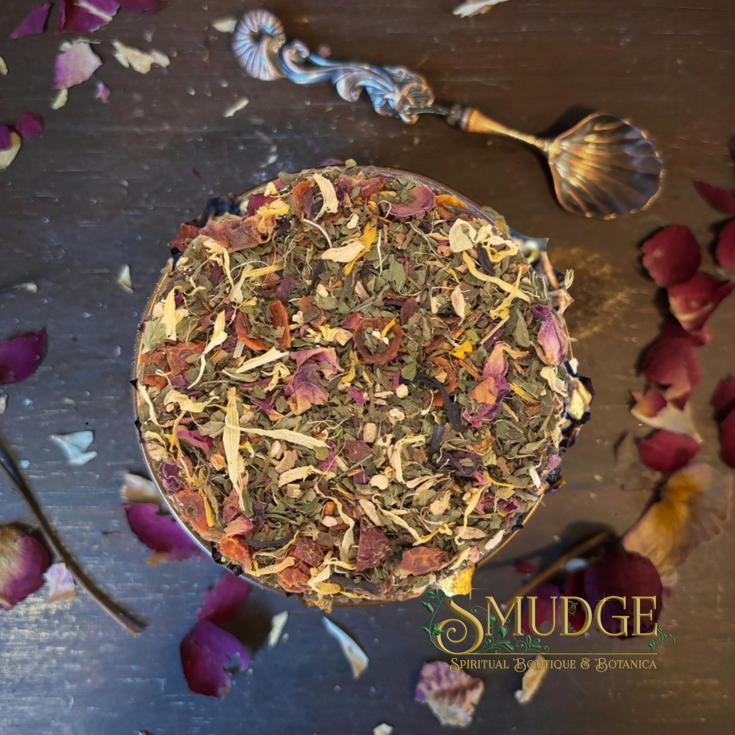 A top-down view of a round tin filled with loose herbal tea blend, featuring dried flowers, leaves, and botanicals, on a dark wooden surface with scattered rose petals and a decorative spoon. The brand logo 'SMUDGE' is visible in the bottom right corner.