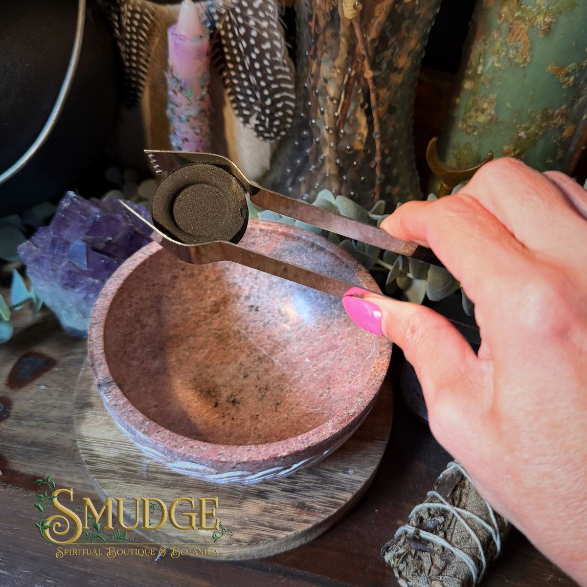 A close-up of a hand with pink nail polish holding a metal scoop over a pinkish-brown stone bowl, with decorative candles, feathers, crystals, and a smudge bundle in a styled setting. The word 'SMUDGE' appears as a logo at the bottom left.