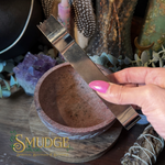A hand with pink nail polish holds a polished metal scraper or spatula above a small terracotta-colored stone bowl, with crystals, dried herbs, and decorative objects in the background. The word 'Smudge' appears as a watermark/logo in the lower left.