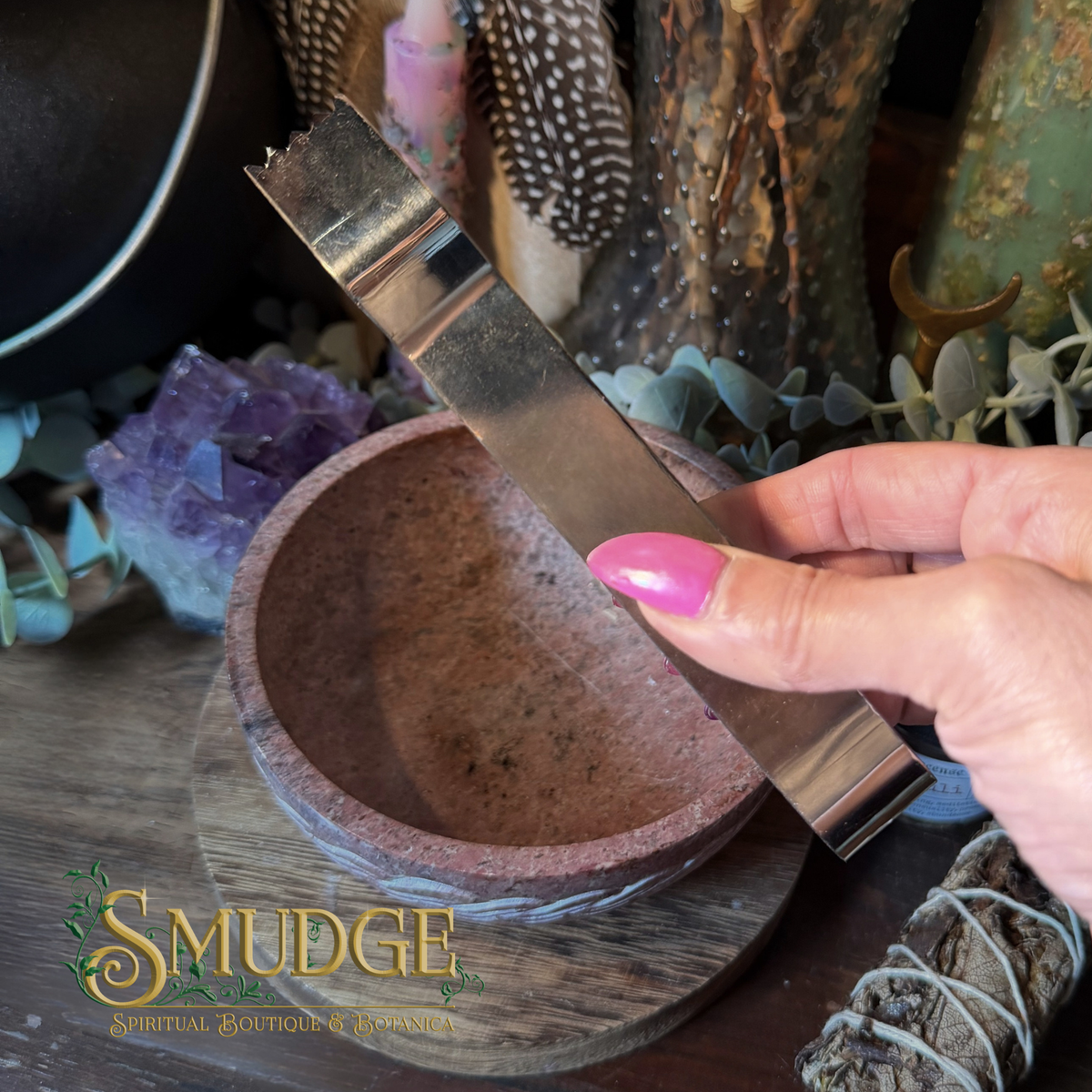 A hand with pink nail polish holds a polished metal scraper or spatula above a small terracotta-colored stone bowl, with crystals, dried herbs, and decorative objects in the background. The word 'Smudge' appears as a watermark/logo in the lower left.
