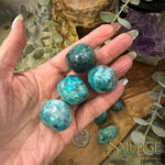A hand holds several polished turquoise-blue jasper stones with white marbling and dark veining, with more stones and decorative botanical props in the background. The Smudge Spirit Shop & Botanica logo appears in the lower right corner.
