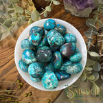 A white ceramic bowl filled with polished turquoise and blue-green gemstone chips, arranged on a wooden surface with eucalyptus leaves and crystals around it. The Smudge Natural Gemstones & Minerals logo is visible in the bottom right corner.