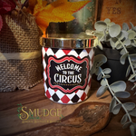 A cylindrical candle tin with a shiny gold lid and a red, black, and white harlequin pattern, featuring a central label that reads 'WELCOME TO THE CIRCUS' in white text. The tin is styled on a rustic wooden surface with autumn leaves, burlap, and greenery in the background.