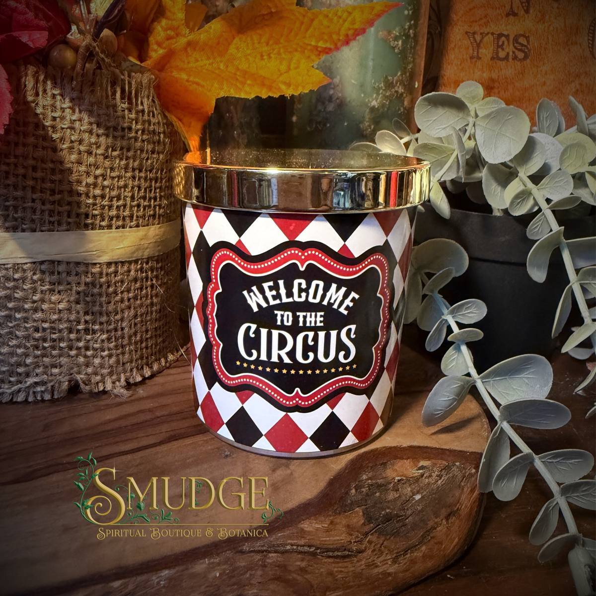 A cylindrical candle tin with a shiny gold lid and a red, black, and white harlequin pattern, featuring a central label that reads 'WELCOME TO THE CIRCUS' in white text. The tin is styled on a rustic wooden surface with autumn leaves, burlap, and greenery in the background.