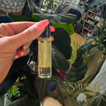 Hand holding a small bottle of liquid with a blurred background featuring plants and decor.