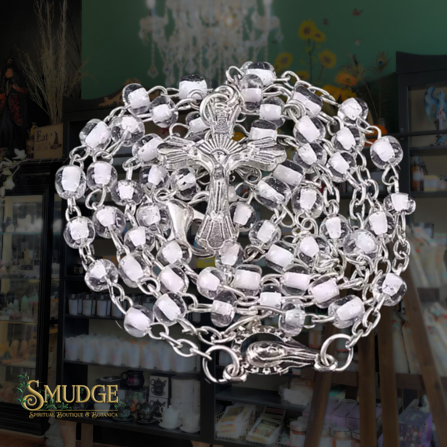 A silver-toned crucifix rosary with clear faceted beads, displayed on a stand inside a shop. The brand logo 'SMUDGE' is visible in the bottom-left corner.