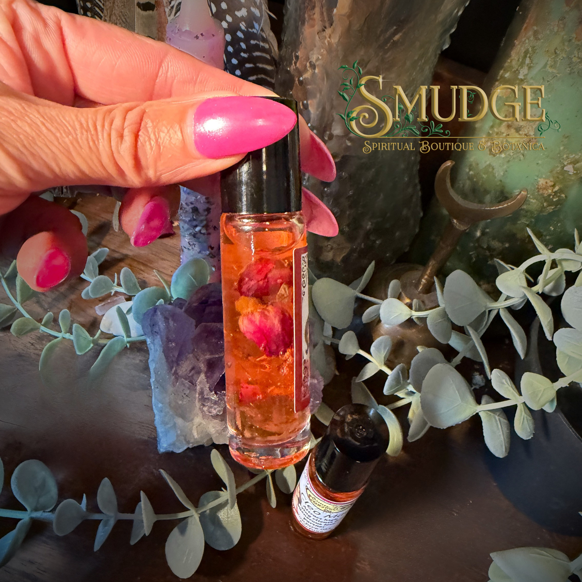 A hand with bright pink nails holds a small glass roller bottle filled with orange liquid and dried flower petals. Another bottle lies nearby on a dark surface surrounded by eucalyptus leaves, crystals, and rustic decor.