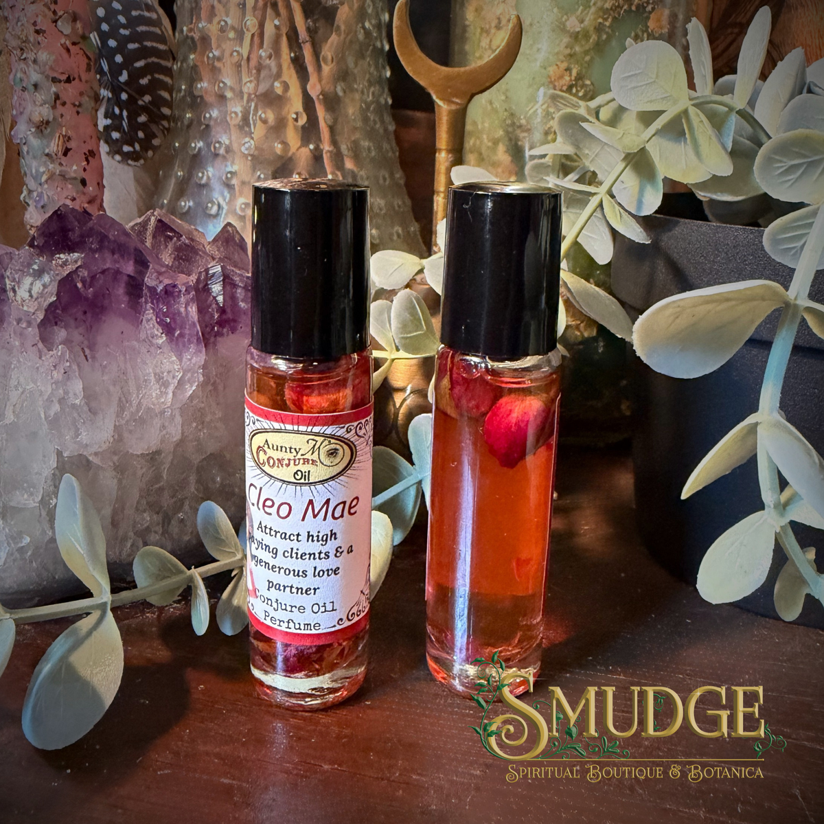 Two small roll-on perfume oil bottles with black caps displayed on a wooden surface, surrounded by decorative crystals, greenery, and a brass spoon. The bottle labels read 'Smudge / Sacred Geometry' and 'Cleo Mae'.