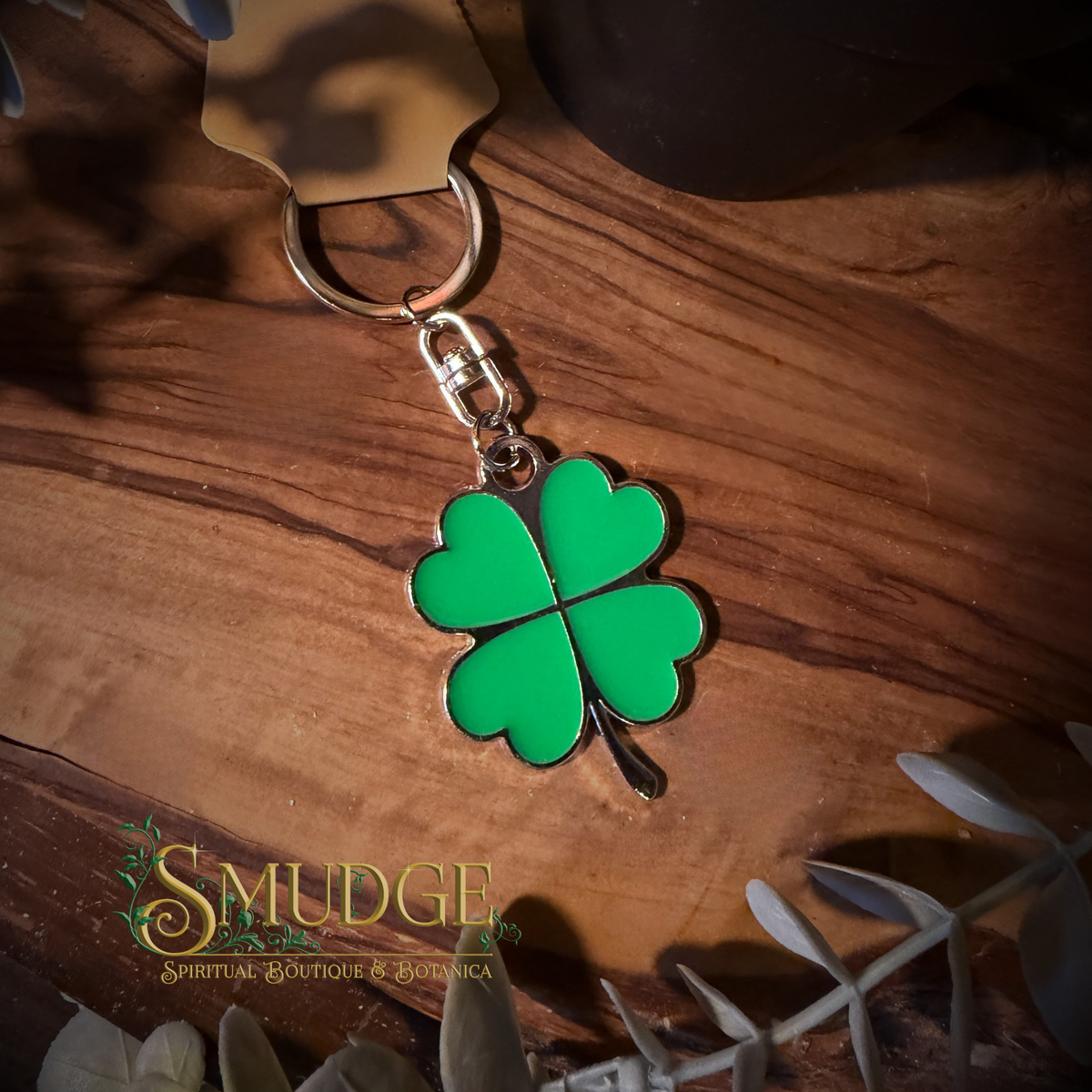 A green enamel four-leaf clover keychain with a silver-tone metal clasp and split ring, resting on a dark wood surface. The brand name 'Smudge Spiritual Boutique & Botanica' is watermarked in the lower-left corner.