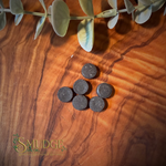 Five small round dark charcoal pads arranged on a warm brown wood surface, with a green leafy stem across the top edge and a visible Smudge logo watermark in the lower left.