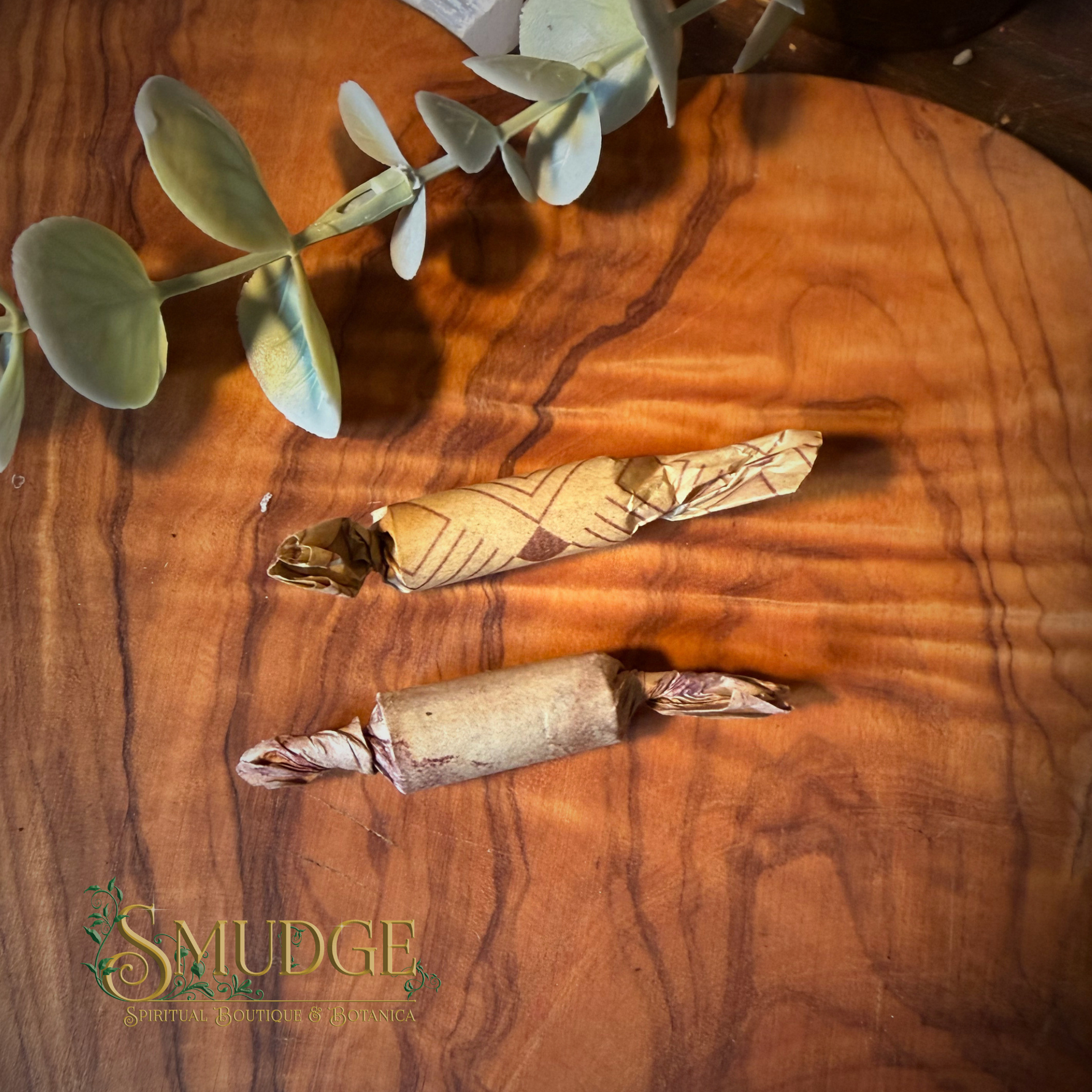 Two wrapped artisanal food items, likely caramel apples or candy-coated treats, wrapped in decorative brown paper, displayed on a polished wooden board with a green eucalyptus sprig and a 'Smudge' brand logo in the corner.