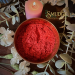 Red incense powder in a bowl with a lit candle and white flowers on a wooden surface, branded 'Smudge'.