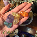 A hand holds several polished tumbled gemstones, including purple, teal, yellow, and orange stones, with a bowl of more stones and greenery in the background. The logo 'SMUDGE' is visible in the lower-left corner.