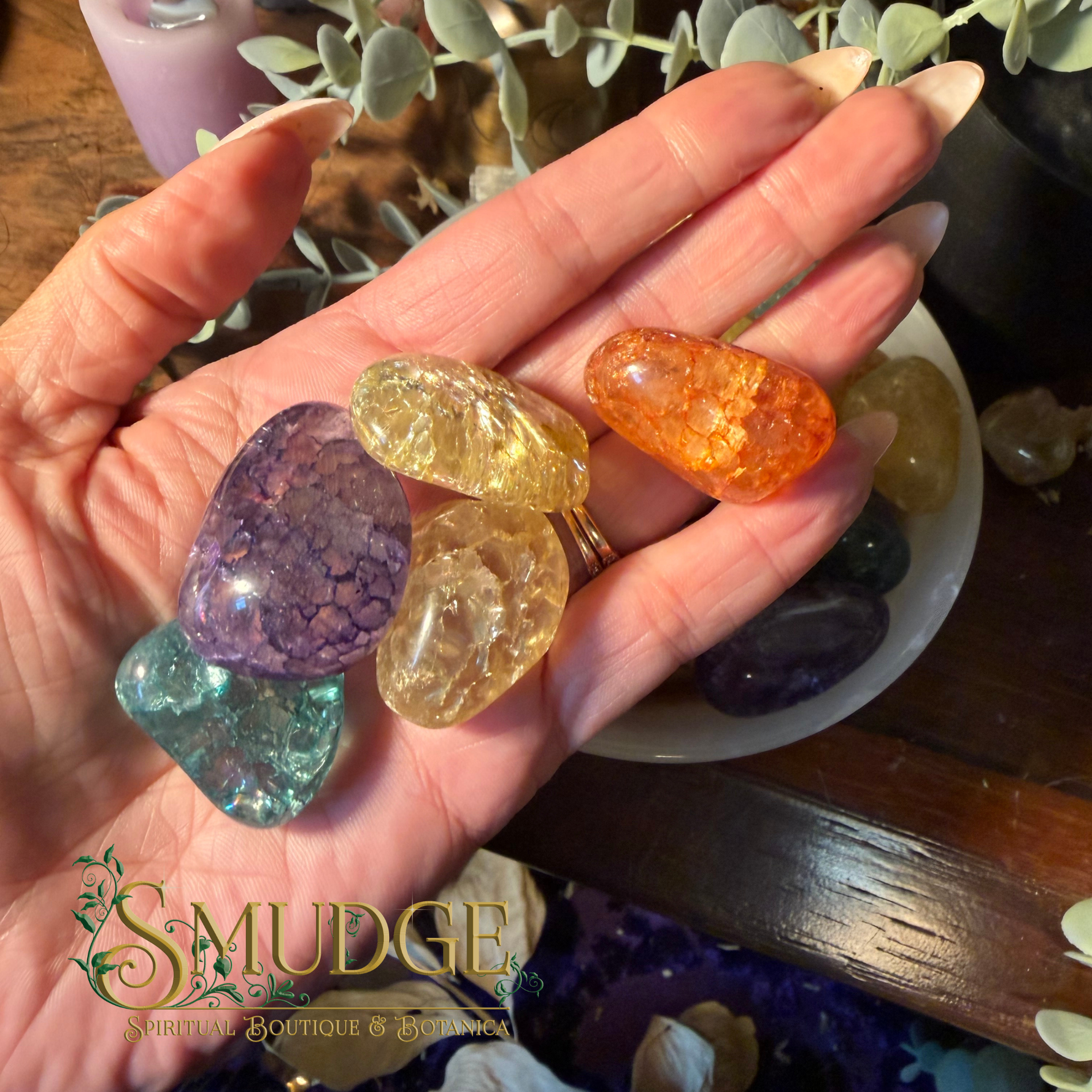 A hand holds several polished tumbled gemstones, including purple, teal, yellow, and orange stones, with a bowl of more stones and greenery in the background. The logo 'SMUDGE' is visible in the lower-left corner.