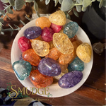 A collection of polished tumbled crystals in various colors, including purple, orange, yellow, and blue, arranged on a white plate. The plate is set on a dark wooden surface with green foliage in the background.