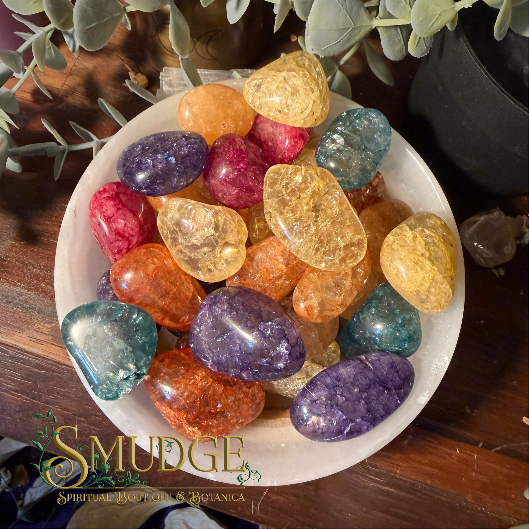 A collection of polished tumbled crystals in various colors, including purple, orange, yellow, and blue, arranged on a white plate. The plate is set on a dark wooden surface with green foliage in the background.