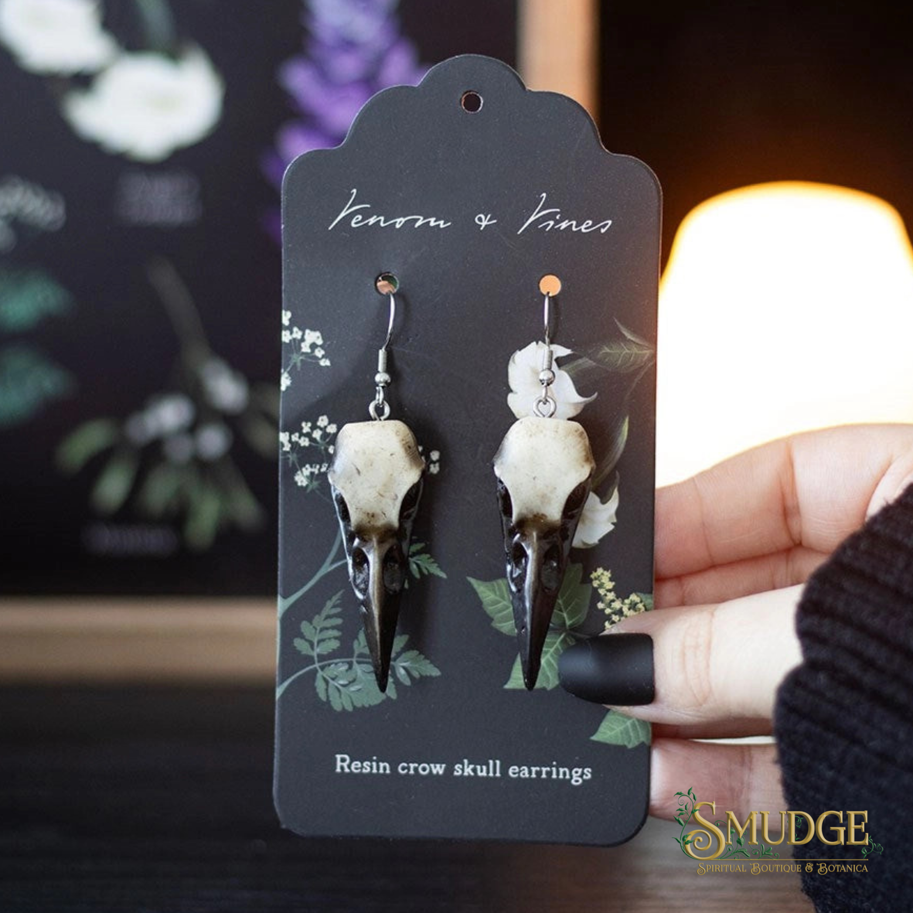 A pair of resin crow skull earrings hanging from a black display card labeled 'Venom & Vines' and 'Resin crow skull earrings.' A hand is holding the card, with a softly blurred indoor background and a warm light source behind it.