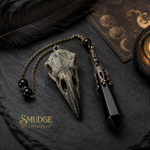 A gothic-style pendant necklace featuring a bird skull and a black crystal point on a dark chain, styled on a dark slate background with a feather, moon phases card, and candle. The brand logo 'SMUDGE Spiritual Jewelry Express' is visible in the bottom-left corner.